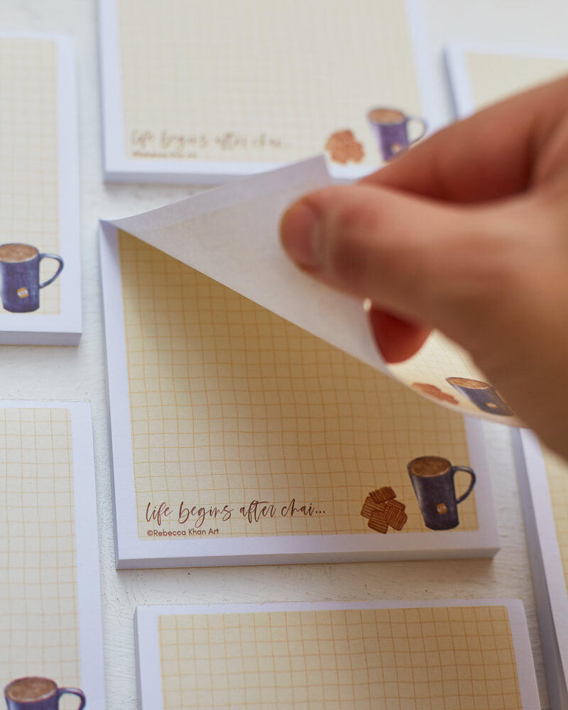 Close up of a hand tearing off a note from a notepad with a pale yellow grid background with a white border, on the bottom a cup of tea and biscuits are depicted with the words "life begins after chai..." in script lettering.
