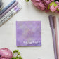 A notepad depicting a starry galaxy against a light purple background with the words "The sky is NOT the limit!!!" in white hand lettering across the bottom.  The notepad are on a desk surrounded by flowers and purple stationery supplies.