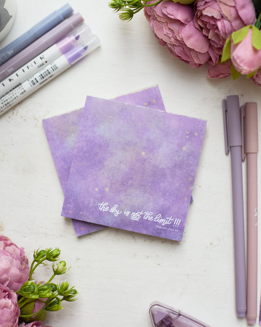 Two notepads depicting a starry galaxy against a light purple background with the words "The sky is NOT the limit!!!" in white hand lettering across the bottom.  The notepads are on a desk surrounded by flowers and purple stationery supplies.