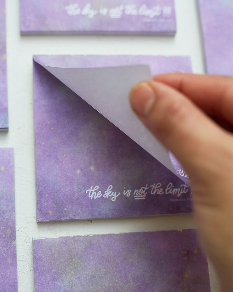Close up of a hand tearing off a note from a notepad depicting a starry galaxy against a light purple background with the words "The sky is NOT the limit!!!" in white hand lettering across the bottom.