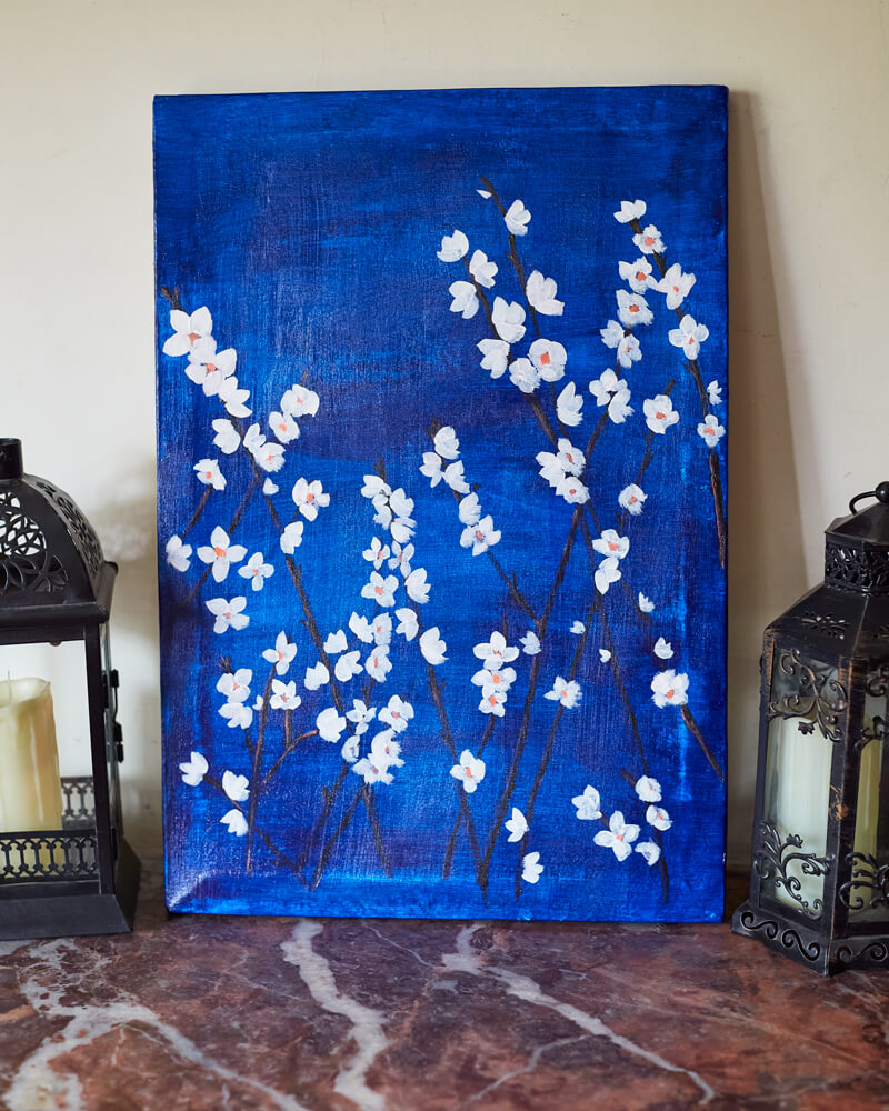 An acrylic painting on canvas depicting white blossoms against a moody blue background.  The painting rests on a warm marble surface flanked by two lanterns.