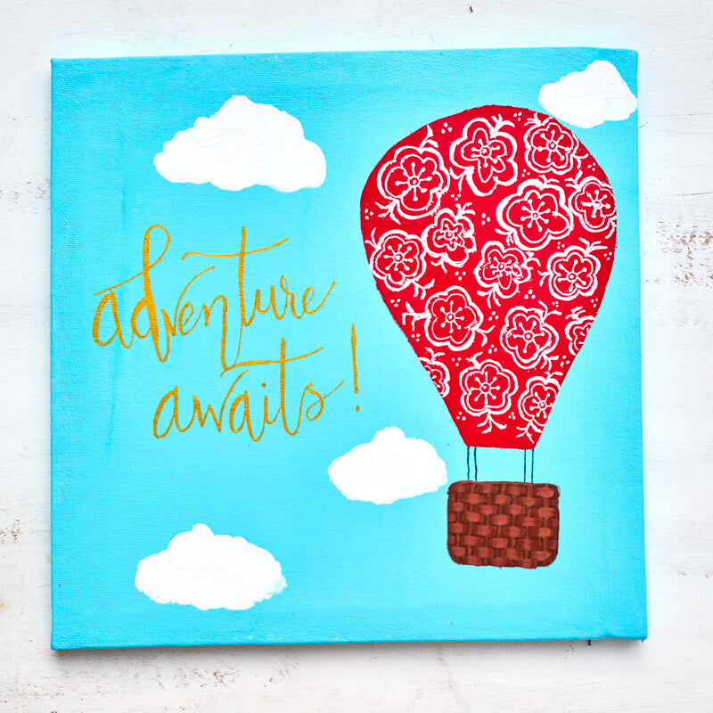 Acrylic painting on canvas of a bright red hot air balloon with hand painted white flowers ascending the sky featuring the words "Adventure Awaits" in metallic gold hand lettering.