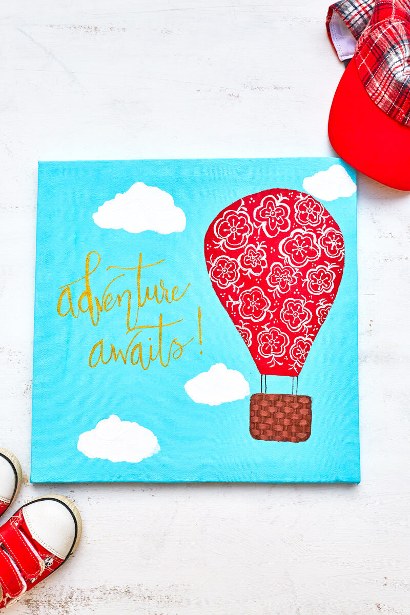 Acrylic painting on canvas of a bright red hot air balloon with hand painted white flowers ascending the sky featuring the words "Adventure Awaits" in metallic gold hand lettering.  The painting is surrounded by a child's red cap and a pair of bright red sneakers.