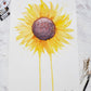 Flat lay of two watercolor tubes, a water color palette, a dried flower and paintbrushes around a watercolor painting of a large sunflower with petals radiating outward and two petals dripping down to the bottom of the painting.