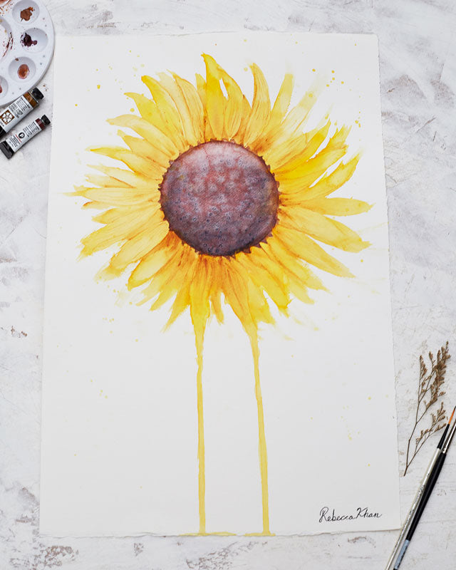 Flat lay of two watercolor tubes, a water color palette, a dried flower and paintbrushes around a watercolor painting of a large sunflower with petals radiating outward and two petals dripping down to the bottom of the painting.