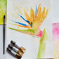 Flat lay of three tubes of watercolor, a brush, some swatches of watercolor and a watercolor painting of a bird of paradise flower painted in a loose, impressionist style.