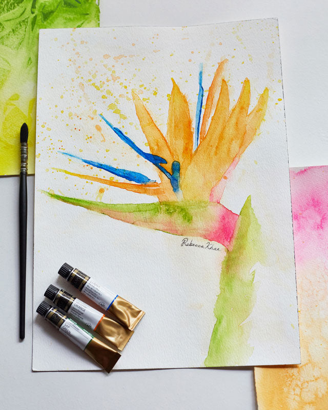 Flat lay of three tubes of watercolor, a brush, some swatches of watercolor and a watercolor painting of a bird of paradise flower painted in a loose, impressionist style.
