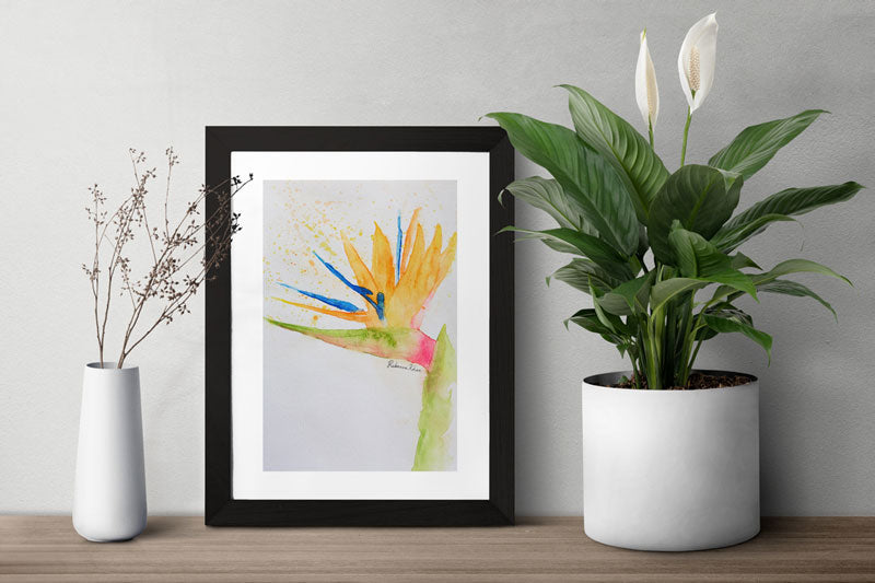 Watercolor painting of a bird of paradise flower painted in a loose, impressionist style in a black frame on a table in between two plants.