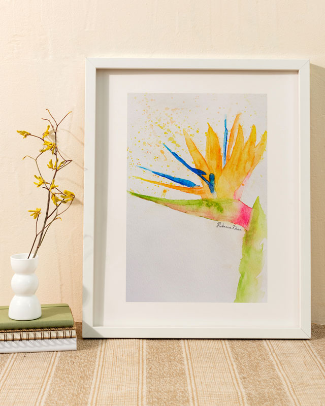 Watercolor painting of a bird of paradise flower painted in a loose, impressionist style in a white frame against a warm colored wall.