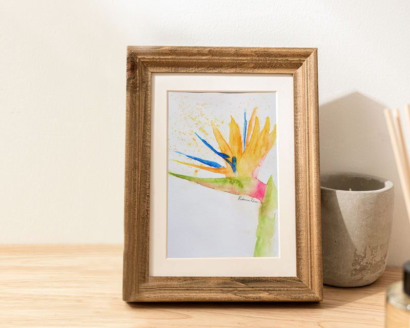 Art print of a watercolor painting of a bird of paradise flower painted in a loose, impressionist style in a wooden frame on a table next to a candle.