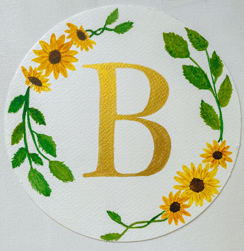 Watercolor monogram of the letter B in gold watercolor surrounded by yellow watercolor sunflowers and green leaves on a round piece of paper.