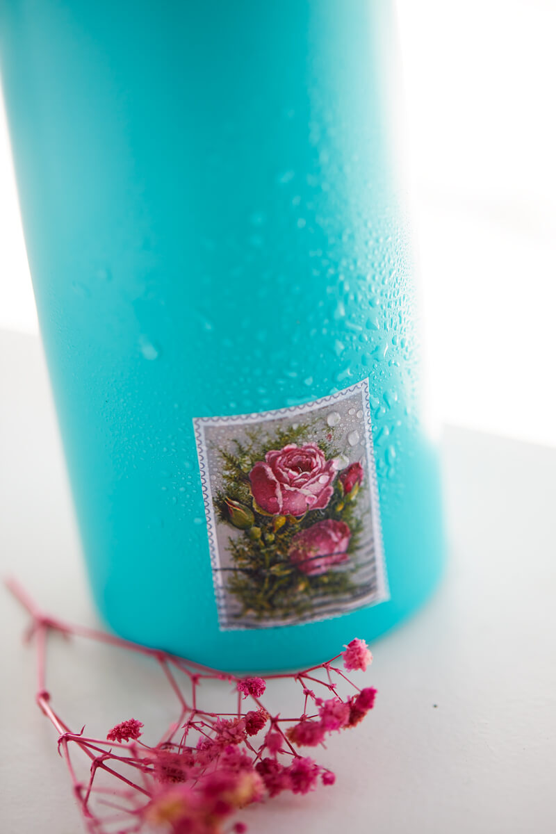 A teal water bottle with a floral vintage stamp sticker sprayed with water to show that it is waterproof.