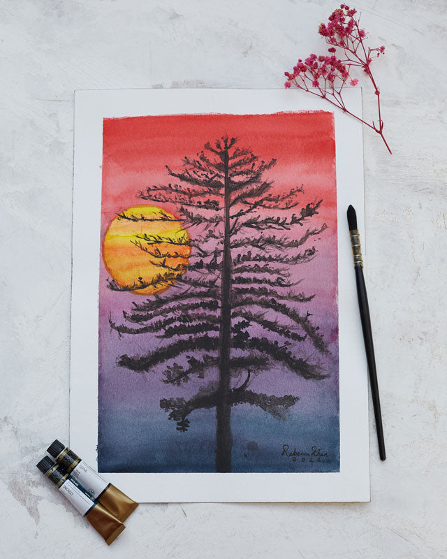 Flat lay of paintbrush, watercolor tubes, a dried flower and a watercolor painting of a thin black tree in front of a sunset with a red sky fading into black.