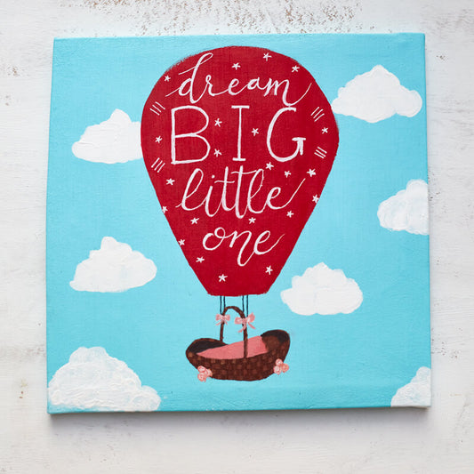 Acrylic painting on canvas of a bright red hot air balloon featuring the words "Dream Big Little One" in hand lettering ascending the sky with a baby basket. 
