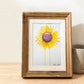 Art print of a watercolor painting of a large sunflower with petals radiating outward and two petals dripping down to the bottom of the painting inside a wooden frame on a table next to a candle.