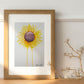 Art print of a watercolor painting of a large sunflower with petals radiating outward and two petals dripping down to the bottom of the painting in a wooden frame on a table with decoration pieces next to it.