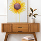 A side table with a plant and a large watercolor painting of a  sunflower with petals radiating outward and two petals dripping down to the bottom of the painting.