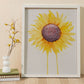 A large watercolor painting of a sunflower with petals radiating outward and two petals dripping down to the bottom of the painting in a white frame against a wall next to books and a vase with dried flowers.