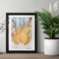A table with two plants and a framed art print of a watercolor painting of two hands holding a bright mango that has been cut into to reveal the inside.