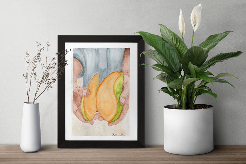 A table with two plants and a framed art print of a watercolor painting of two hands holding a bright mango that has been cut into to reveal the inside.