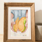 A wooden frame with an art print of a watercolor painting of two hands holding a bright mango that has been cut into to reveal the inside.