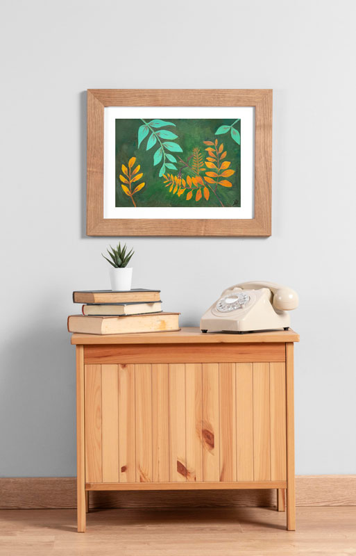 A small side table with books, a plant, a telephone, and above it, hanging on the wall in a wooden frame is an art print of a gouache painting of gold and teal leaves reaching towards each other against a dark green background.