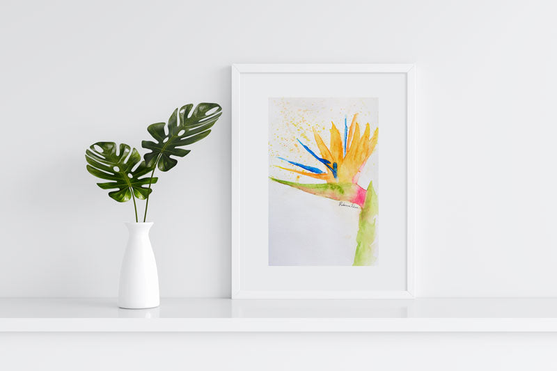 Watercolor painting of a bird of paradise flower painted in a loose, impressionist style in a white frame on a white shelf against a white wall next to a white vase with two leaves.