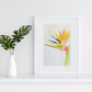 Art print of a watercolor painting of a bird of paradise flower painted in a loose, impressionist style in a white frame on a white shelf against a white wall next to a white vase with two leaves.