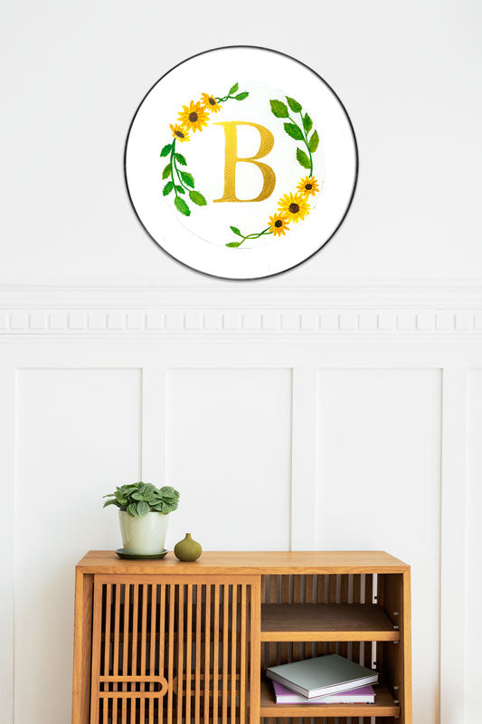 Watercolor monogram of the letter B in gold watercolor surrounded by yellow watercolor sunflowers and green leaves on a round piece of paper in a round frame above a side table.