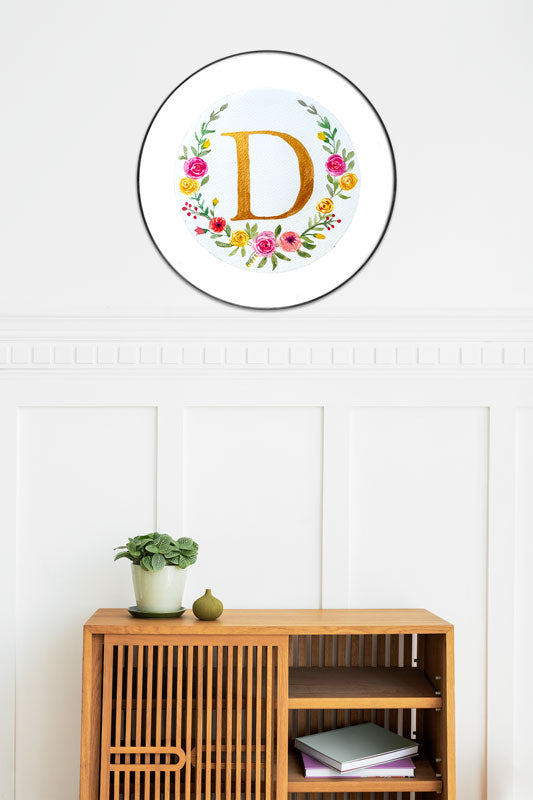 Watercolor monogram of the letter D in gold watercolor surrounded by pink and yellow watercolor flowers and green leaves on a round piece of paper in a round frame above a side table.