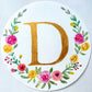 Watercolor monogram of the letter D in gold watercolor surrounded by pink and yellow watercolor flowers and green leaves on a round piece of paper.