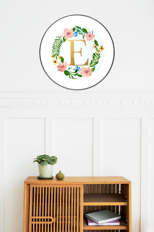 Watercolor monogram of the letter E in gold watercolor surrounded by orange watercolor hibiscus flowers and green leaves on a round piece of paper in a round frame above a side table.
