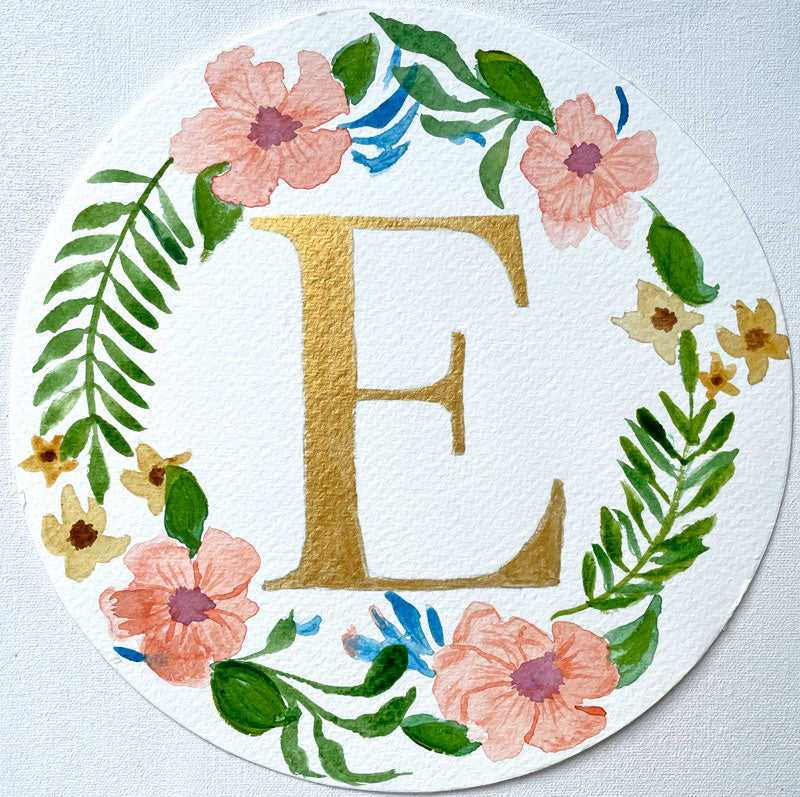 Watercolor monogram of the letter E in gold watercolor surrounded by orange watercolor hibiscus flowers and green leaves on a round piece of paper.