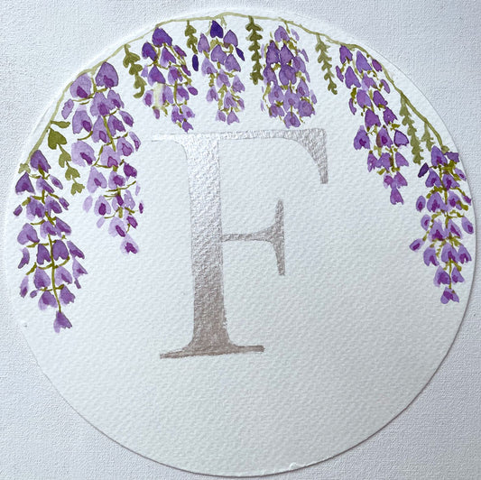Watercolor monogram of the letter F in silver watercolor with purple watercolor wisteria hanging down around it on a round piece of paper.