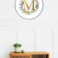 Watercolor monogram of the letter M in gold watercolor surrounded by pink and peach watercolor peonies and green and brown leaves on a round piece of paper in a round frame above a side table.