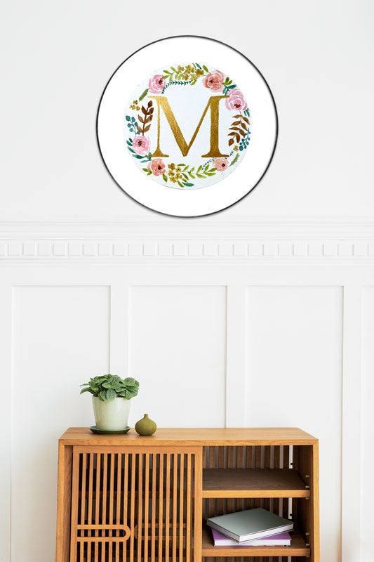 Watercolor monogram of the letter M in gold watercolor surrounded by pink and peach watercolor peonies and green and brown leaves on a round piece of paper in a round frame above a side table.