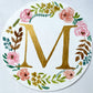 Watercolor monogram of the letter M in gold watercolor surrounded by pink and peach watercolor peonies and green and brown leaves on a round piece of paper.