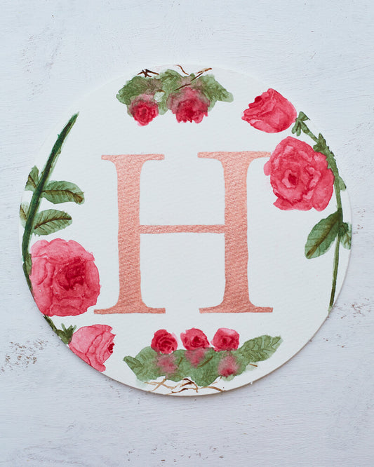 Round watercolor monogram of the letter H written in a metallic rose paint surrounded by watercolor roses.