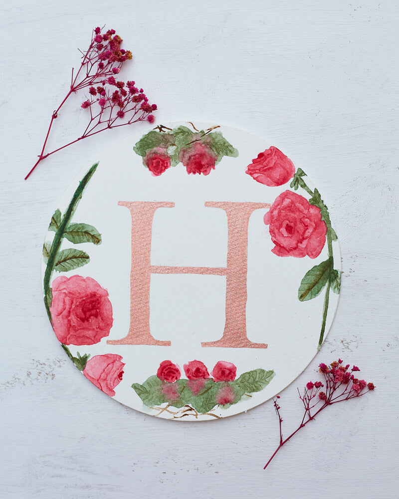 Two dried red flowers flanking a round watercolor monogram of the letter H written in a metallic rose paint surrounded by watercolor roses.