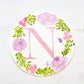 Round watercolor monogram painting of the letter N painted in duochrome pink and purple metallic paint surrounded by painted purple peonies and pink roses and green leaves.