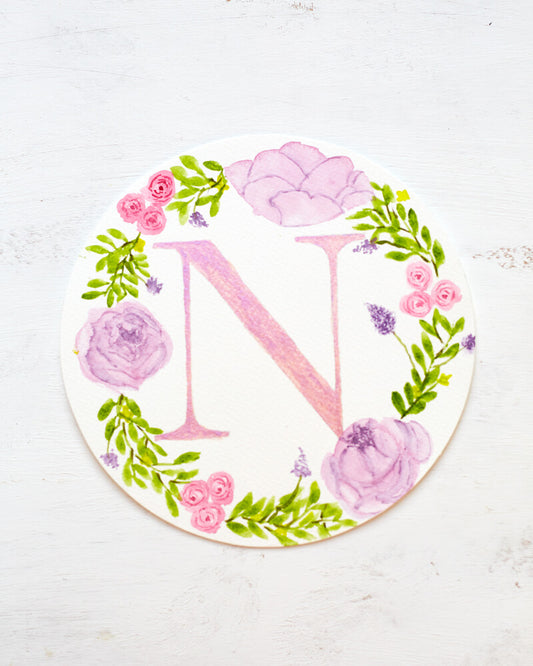 Round watercolor monogram painting of the letter N painted in duochrome pink and purple metallic paint surrounded by painted purple peonies and pink roses and green leaves.