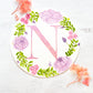 Round watercolor monogram painting of the letter N painted in duochrome pink and purple metallic paint surrounded by painted purple peonies and pink roses and green leaves flanked by dried pink flowers.