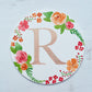 Watercolor monogram of the letter R painted in metallic duochrome peach and green paint with red and orange flowers and green leaves painted around it on a round paper.