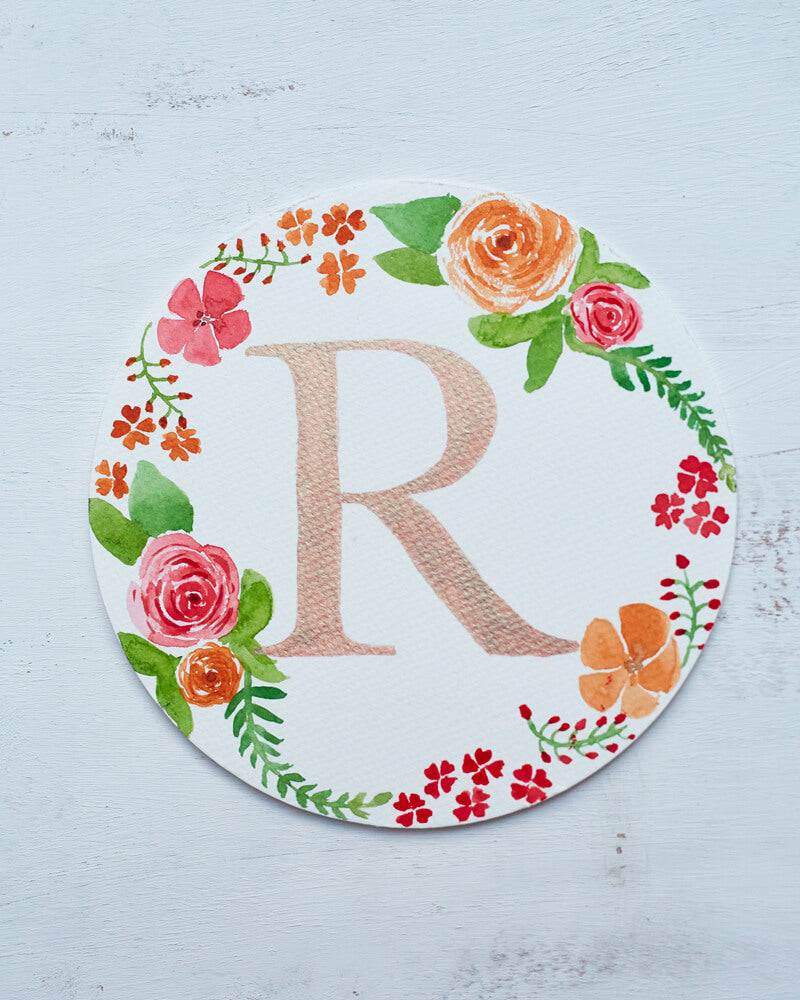 Watercolor monogram of the letter R painted in metallic duochrome peach and green paint with red and orange flowers and green leaves painted around it on a round paper.