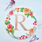 Two dried red flowers flanking a watercolor monogram of the letter R painted in metallic duochrome peach and green paint with red and orange flowers and green leaves painted around it on a round paper.