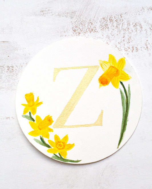 Watercolor monogram of the letter Z painted in duochrome metallic paint with pale gold and green shine surrounded by painted daffodils on a round piece of paper.