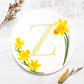 Two dried flowers flanking a watercolor monogram of the letter Z painted in duochrome metallic paint with pale gold and green shine surrounded by painted daffodils on a round piece of paper.