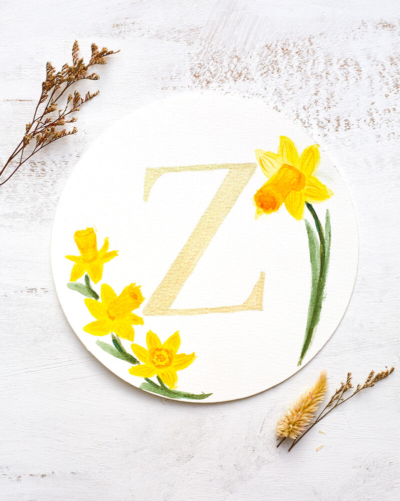 Two dried flowers flanking a watercolor monogram of the letter Z painted in duochrome metallic paint with pale gold and green shine surrounded by painted daffodils on a round piece of paper.
