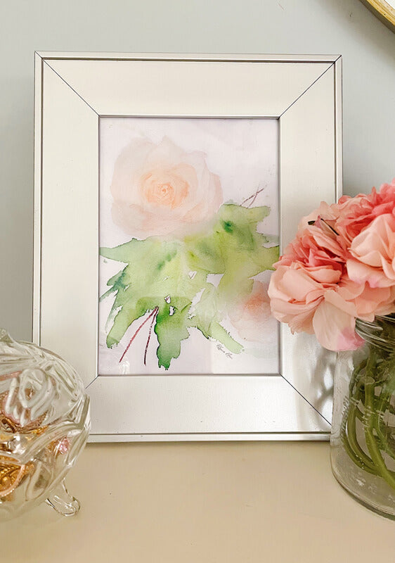 An art print of a peach rose painted with watercolors in a loose, impressionist style inside of a silvr frame on a table next to a glass candy dish and a glass vase of pink flowers..