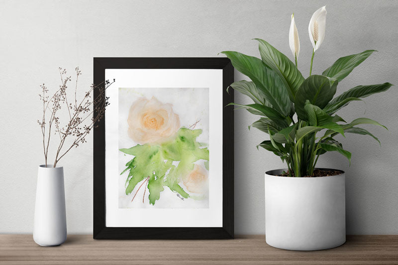 Framed art print of a peach rose painted with watercolors in a loose, impressionist style on a table in between two plants.